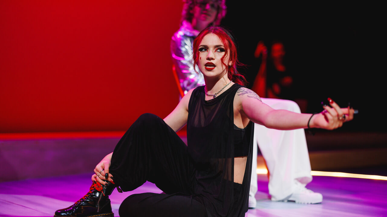 A female student actor on stage with red background lighting for the productions, Thebes