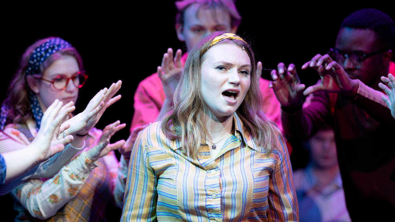 A female student actor surrounded by other student actors singing on stage for the production, Rage